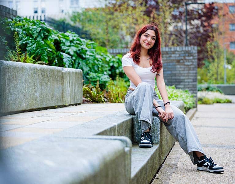 A student sat on some steps outdoors with shrubbery behind her, smiling at the camera.
