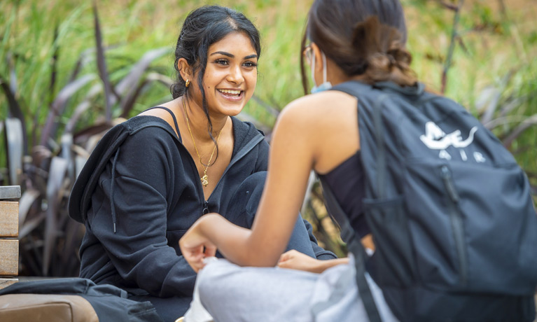 Two international student wearing backpacks, sitting and talking outside on campus with greenery and trees behind them.