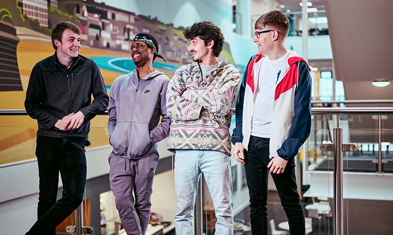 Four students laughing together leaning against an indoor balcony.