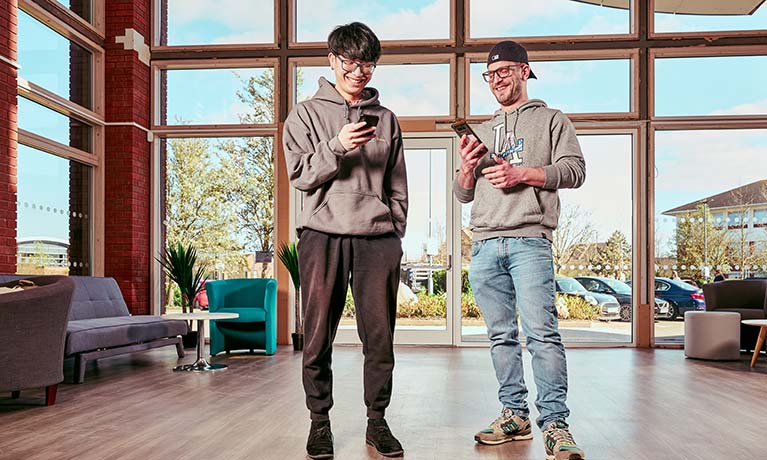 Two students stood in an open area looking at their phones