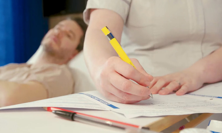 A student nurse writing down notes with a patient in a bed behind them.